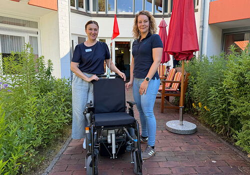 Zwei Frauen stehen neben einem Rollstuhl im Freien.