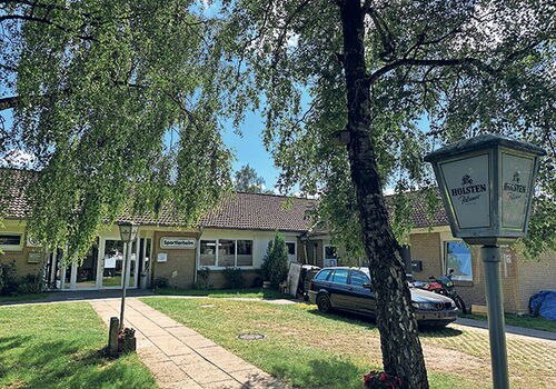 Sportlerheim mit Bäumen und parkendem Auto bei Sonnenschein.