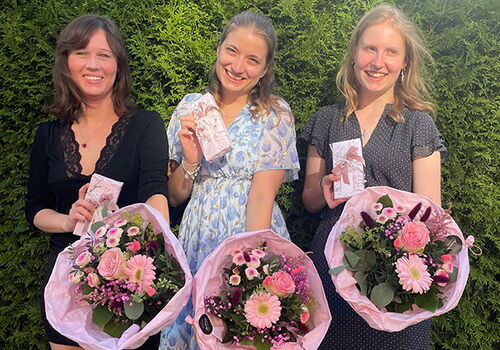 Drei Frauen mit Blumensträußen und Karten lächeln vor einer Hecke.