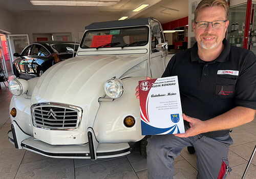 Mann hält Zertifikat neben einem weißen Citroën 2CV in der Werkstatt.