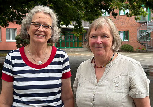 Zwei Frauen lächeln im Freien, Bäume und Gebäude im Hintergrund.