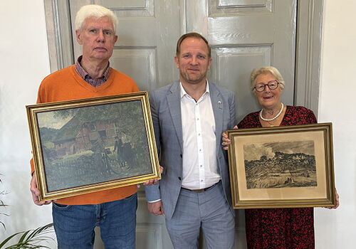Drei Personen halten gerahmte Bilder vor einer grauen Tür.