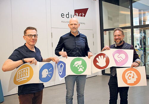 Drei Männer halten Plakate mit bunten Symbolen vor edulab-Tür.