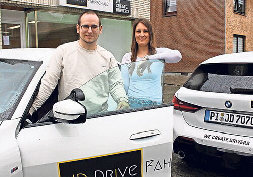 Zwei Personen vor einer Fahrschule mit Autos im Hintergrund.
