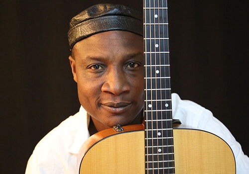 Mann mit Gitarre schaut direkt in die Kamera, trägt Lederhut.