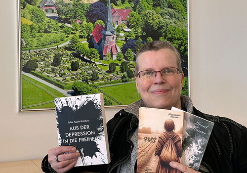 Person mit Brille hält drei Bücher vor Luftbild.