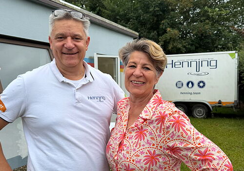 Zwei Personen lächeln vor einem Gebäude mit Henning-Logo.