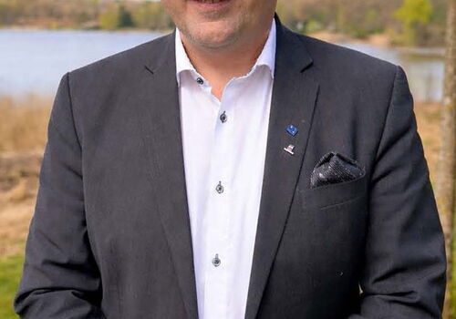 Mann im Anzug lächelt im Freien vor einem See.