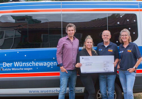 Vier Personen halten einen großen Scheck vor einem Wünschewagen.