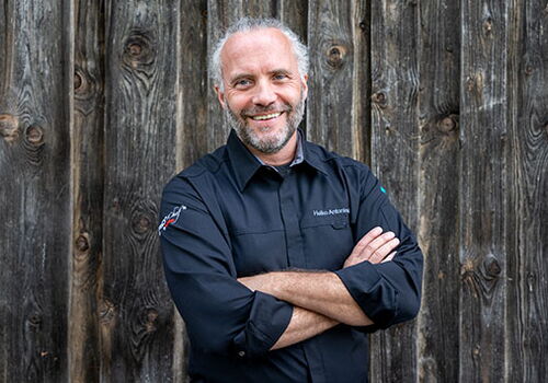 Mann lächelt vor einer Holzwand mit verschränkten Armen.