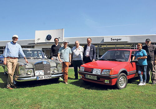 Gruppe posiert mit Oldtimern vor Autohaus.
