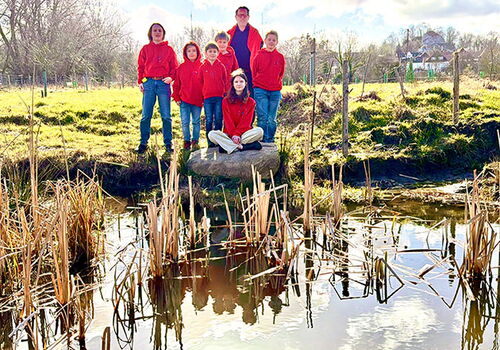 Gruppe steht am Teichrand, sonniger Tag, Schilf im Vordergrund.