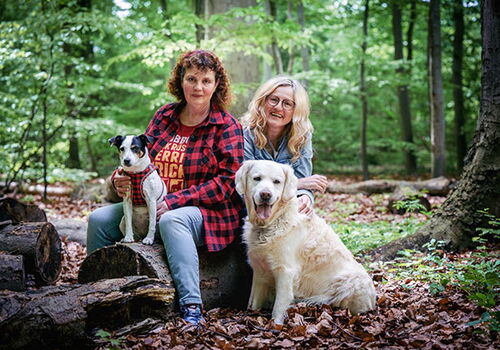 Zwei Frauen mit zwei Hunden im Wald auf Baumstämmen sitzend.