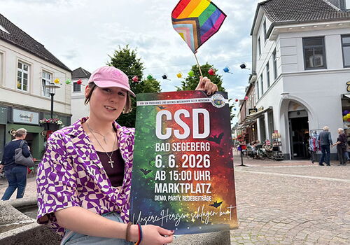 Person hält CSD-Plakat auf einem Stadtplatz.