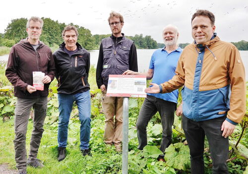 Fünf Männer stehen draußen um ein Schild am See.