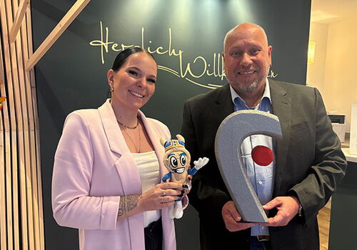 Zwei Personen mit einem Maskottchen und einer Trophäe in der Hand.