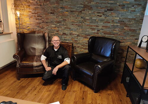 Mann kniet zwischen zwei Sesseln vor einer Ziegelmauer.