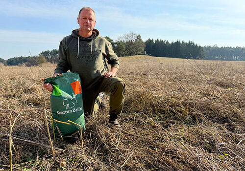 Mann kniet auf trockenem Feld mit Saatgutbeutel.