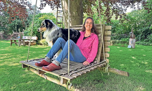 Frau sitzt mit Hund auf einer Holzschaukel im Garten.