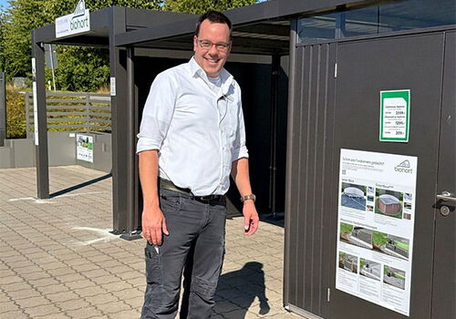 Mann in weißem Hemd vor einem Gartenhaus auf einem Parkplatz
