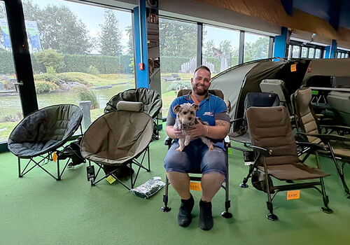 Mann mit Hund auf Stuhl in Campingladen, Zelte im Hintergrund.