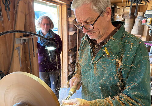 Mann drechselt Holz in der Werkstatt, eine Person schaut zu.