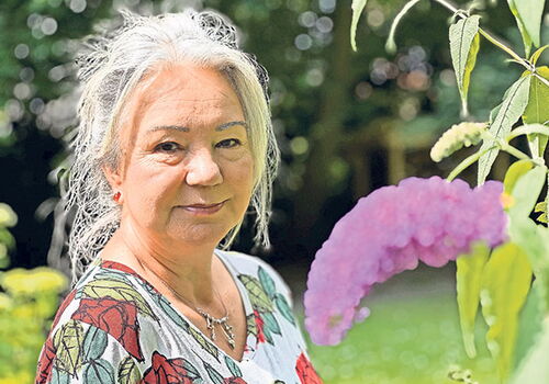 Ältere Frau im Garten mit lila Blüte im Vordergrund.
