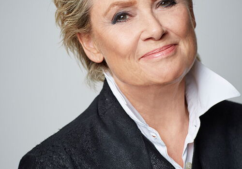 Porträt einer Person mit kurzem, lockigem Haar und einem schwarzen Blazer.