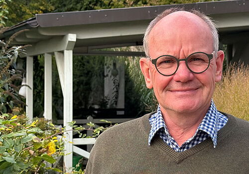 Mann mit Brille vor Gartenhaus und Pflanzen.