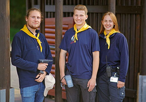 Drei junge Erwachsene mit gelben Halstüchern lächeln in die Kamera.