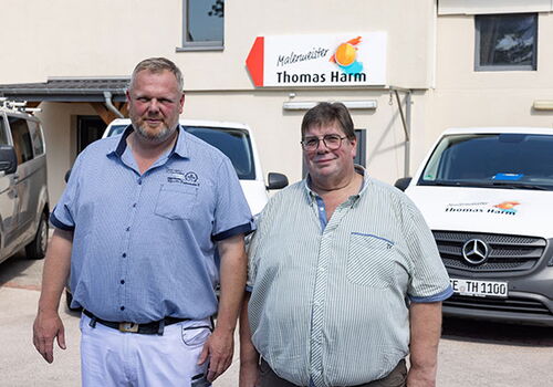 Zwei Männer vor einem Malergeschäft mit Autos im Hintergrund.