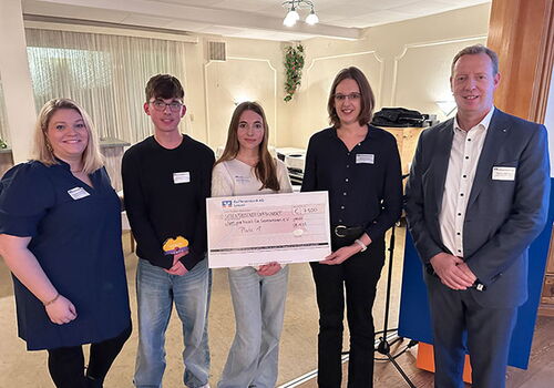 Fünf Personen halten einen großen Scheck in einem Raum.