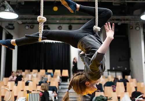 Mädchen balanciert kopfüber an Seil in Theaterumgebung.