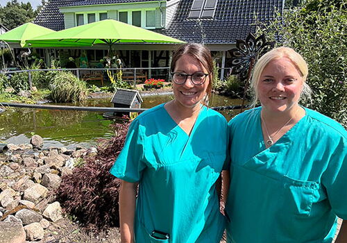 Zwei Krankenschwestern vor einem Gartenteich mit Haus im Hintergrund.