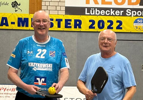 Zwei Männer mit Pickleball-Schlägern und Bällen vor einem Netz.