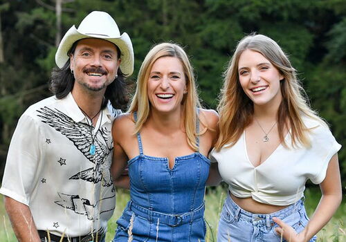 Drei Personen lachen auf einer Wiese in sommerlicher Kleidung.