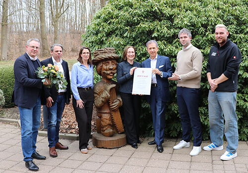 Gruppe posiert mit Zertifikat neben Holzfigur im Freien.