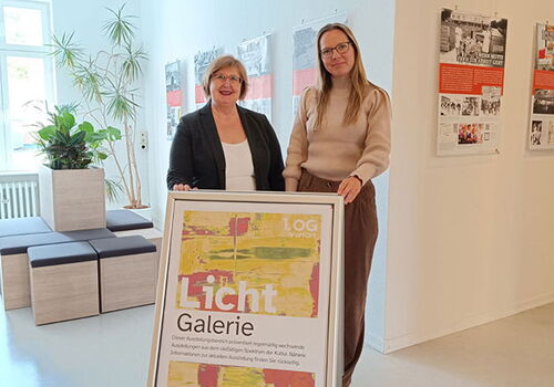 Zwei Frauen posieren neben einem Galerieschild in einem Ausstellungsraum.