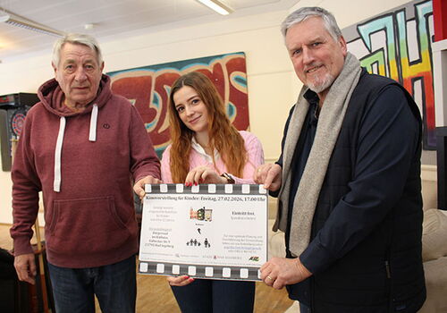 Drei Personen halten ein großes Poster mit Veranstaltungsdetails.