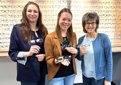 Drei Frauen zeigen verschiedene Brillen vor Brillenregal.