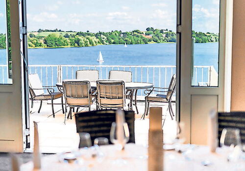 Blick von Terrasse auf See mit Segelboot und Bäumen.