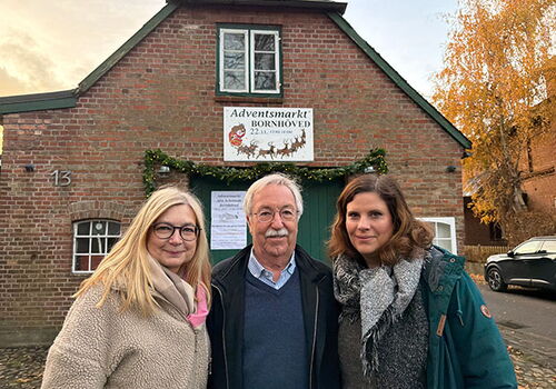 Drei Personen vor einem Haus mit Adventsmarkt-Schild.