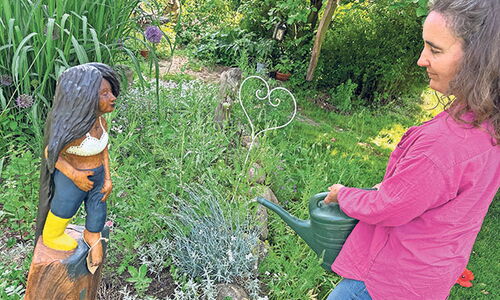 Frau gießt Pflanzen im Garten neben Holzstatue.