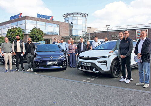 Gruppe posiert neben zwei Autos vor Gebäude.