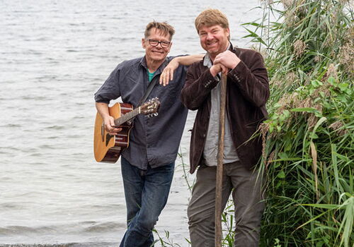 Zwei Männer am Seeufer, einer mit Gitarre.