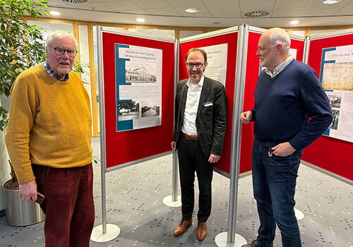 Drei Männer vor rot hinterlegten Infotafeln in einem Raum.