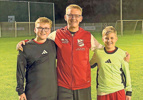 Drei Personen stehen auf einem Fußballplatz und lächeln.
