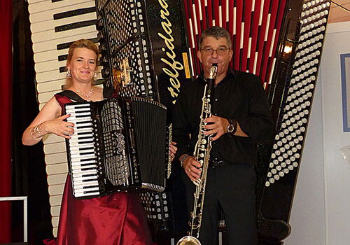 Frau mit Akkordeon und Mann mit Saxophon vor riesigem Akkordeon