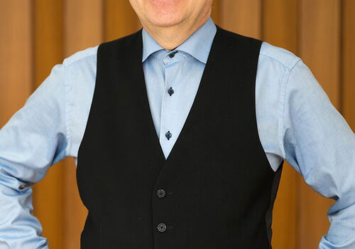 Mann in Hemd und Weste lächelt vor Holzwand.