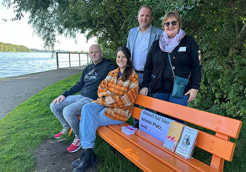 Vier Personen sitzen und stehen neben einer orangefarbenen Bank am Wasser.